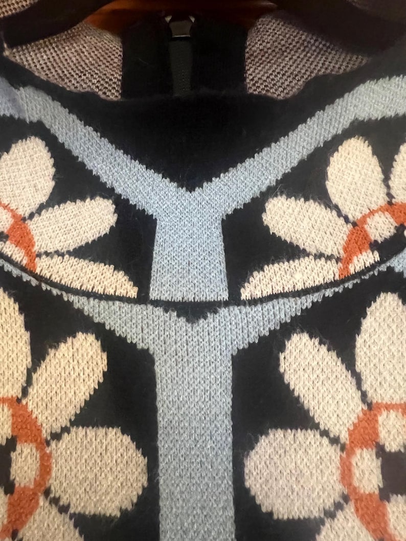 May include: Close-up of a black sweater with a floral pattern. The design features large white and orange flowers with light blue accents. The sweater has a zipper at the neckline and a textured knit.
