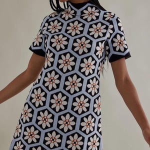 May include: A short-sleeved dress with a high neckline. The dress features a repeating floral pattern in white, orange, and black, set against a light blue hexagonal background. The sleeves and neckline are trimmed in black.