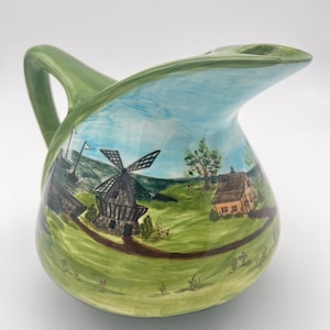 May include: Green ceramic pitcher with a painted scene of a windmill, houses, and trees. The pitcher has a wide spout and a handle.