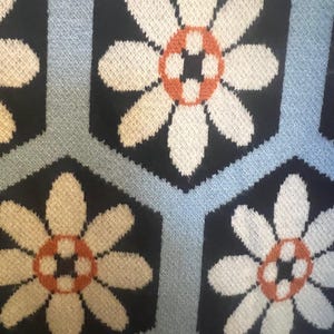 May include: Close-up of a knitted fabric with a repeating floral pattern. The flowers have white petals and orange and white centers, set against a black hexagonal background. Light blue lines separate the hexagons.