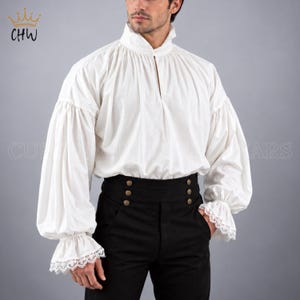 Op de afbeelding: Een wit overhemd met lange mouwen, een hoge kraag, geplooide stof en manchetten met kant, gecombineerd met een zwarte broek met hoge taille en twee verticale rijen messingkleurige knopen. Het overhemd heeft een split bij de hals.