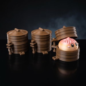 May include: Three brown cylindrical containers with lids, designed to resemble bamboo steamers. Two are closed, while the third is open, revealing a small, round, peach-colored bun with a smiling face, black eyes, and pink cheeks inside.