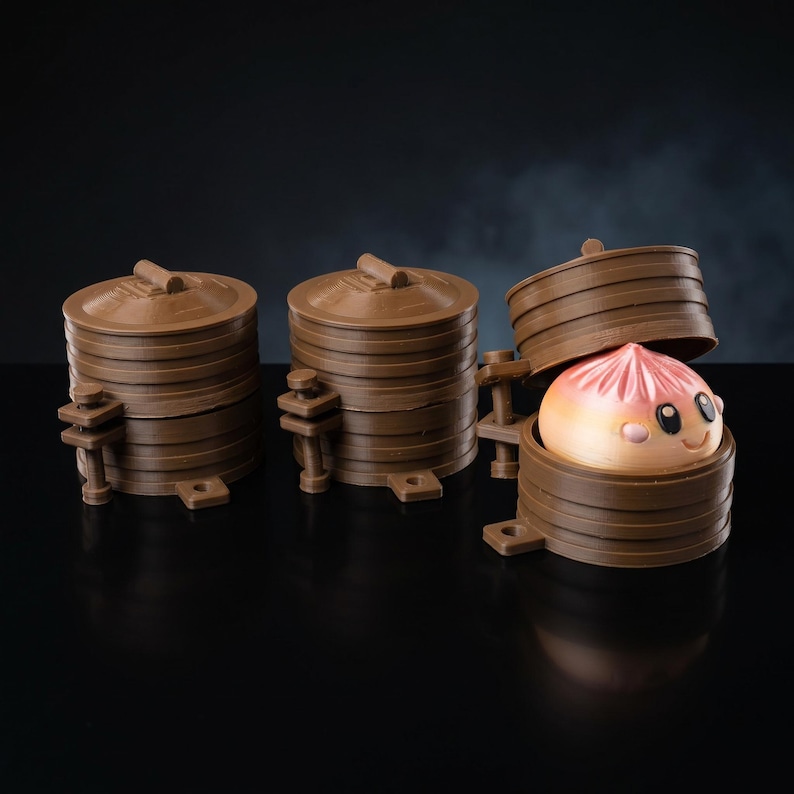 May include: Three brown cylindrical containers with lids, designed to resemble bamboo steamers. Two are closed, while the third is open, revealing a small, round, peach-colored bun with a smiling face, black eyes, and pink cheeks inside.