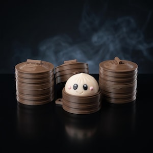 May include: A small, round, cream-colored dumpling figurine with a smiling face, black eyes, and pink cheeks, nestled inside a brown, cylindrical, stackable container. Two additional brown, lidded, stackable containers stand on either side.