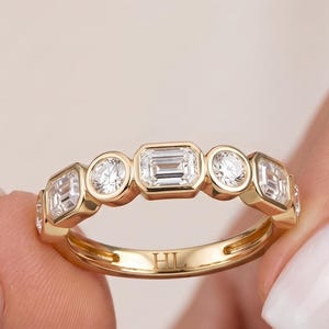 May include: A gold band ring featuring a series of alternating emerald-cut and round-cut clear stones set in a bezel style. The interior of the band is engraved with the letters HL. The stones are arranged horizontally across the top of the ring.