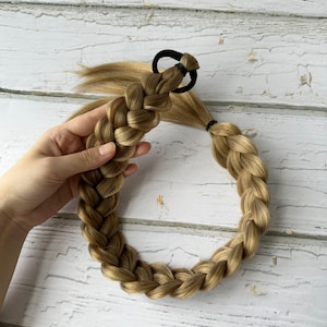 May include: A synthetic hair extension styled into a thick, three-strand braid with a golden blonde color palette. The braid is secured at the top with a black elastic hair tie and features loose, straight hair strands extending from the base.