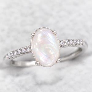 Op de afbeelding: Een zilverkleurige ring met een grote, ovaalvormige, iriserende witte cabochonsteen die door vier pootjes wordt vastgehouden. De ringband is gemaakt van een metaalachtig materiaal en is aan beide zijden bezet met kleine, heldere, ronde steentjes.