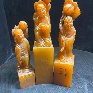 Peut inclure: Trois sculptures en pierre de couleur ambre représentant des personnages, chacune montée sur une base rectangulaire. Les personnages ont le crâne rasé, portent des robes et tiennent des éléments en forme de lotus. Chaque base porte des inscriptions.