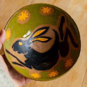 May include: A round ceramic bowl with a glossy olive green glaze featuring a hand-painted black rabbit with yellow ear accents. The bowl is decorated with several small, bright yellow star-shaped motifs scattered across the green surface.