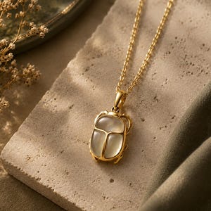 Puede incluir: Un collar de tono dorado con un colgante de escarabajo. El colgante tiene un marco de metal dorado pulido y una incrustaci&oacute;n de piedra ovalada transl&uacute;cida de color blanco lechoso. La cadena es de estilo de eslabones finos en tono dorado.