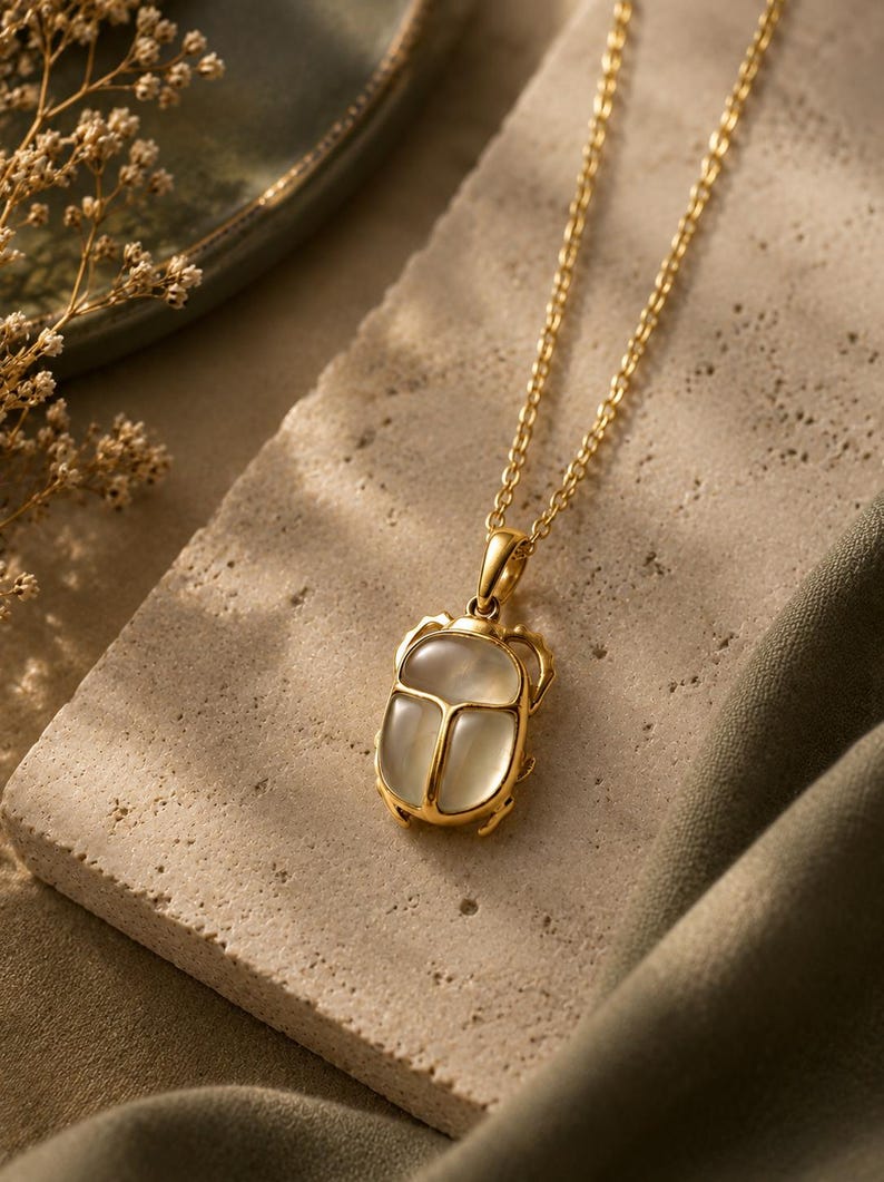 Puede incluir: Un collar de tono dorado con un colgante de escarabajo. El colgante tiene un marco de metal dorado pulido y una incrustaci&oacute;n de piedra ovalada transl&uacute;cida de color blanco lechoso. La cadena es de estilo de eslabones finos en tono dorado.