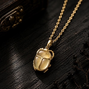 May include: A gold-toned necklace featuring a scarab beetle pendant with a smooth, translucent, pale yellow stone inlay. The pendant is attached to a delicate gold-toned chain, showcasing a detailed metallic beetle frame with visible legs.