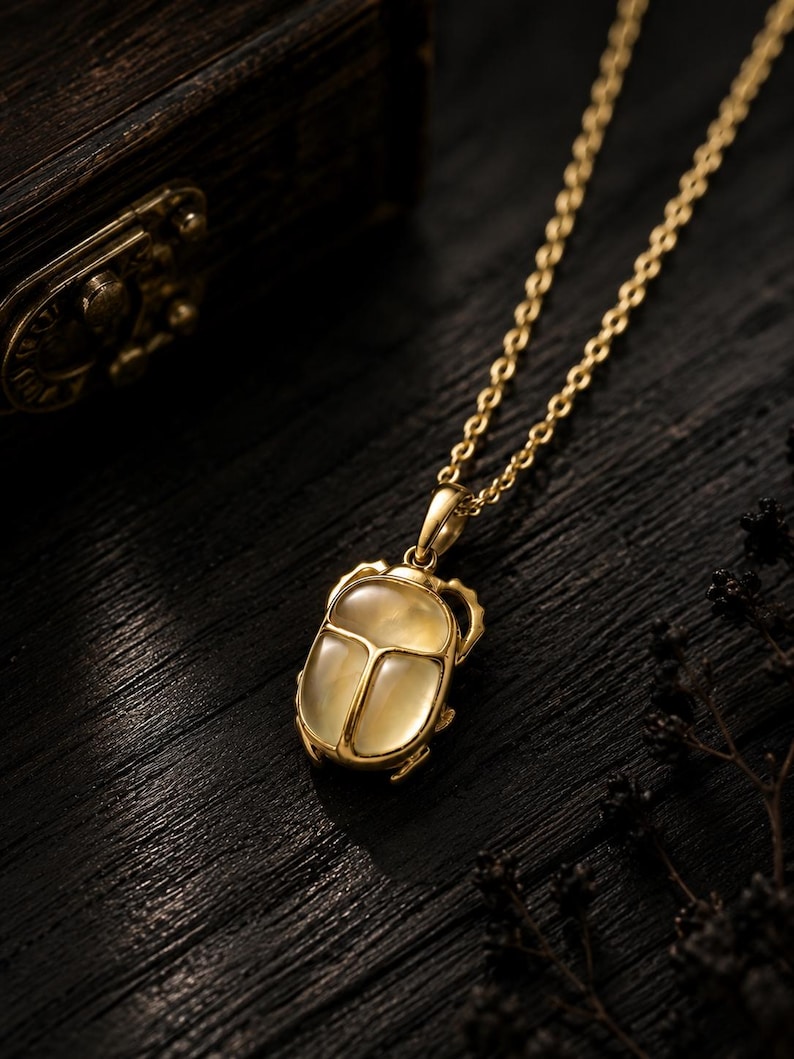 Puede incluir: Un collar de tono dorado con un colgante de escarabajo que presenta una incrustaci&oacute;n de piedra lisa, transl&uacute;cida y de color amarillo p&aacute;lido. El colgante est&aacute; unido a una delicada cadena dorada, mostrando un marco met&aacute;lico con patas visibles.