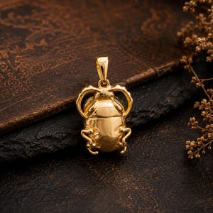 Puede incluir: Un colgante de tono dorado pulido con forma de escarabajo, que presenta un cuerpo liso y redondeado, patas curvas y una peque&ntilde;a anilla integrada en la parte superior para sujetarlo a una cadena. La superficie met&aacute;lica refleja la luz.