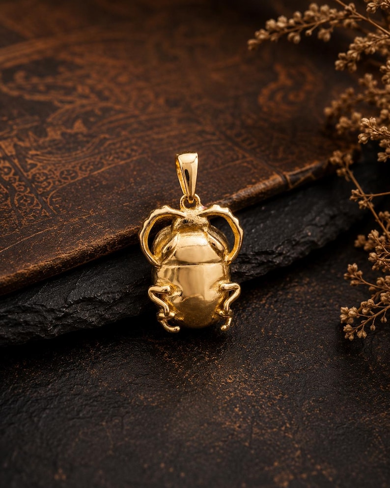 Puede incluir: Un colgante de tono dorado pulido con forma de escarabajo, que presenta un cuerpo liso y redondeado, patas curvas y una peque&ntilde;a anilla integrada en la parte superior para sujetarlo a una cadena. La superficie met&aacute;lica refleja la luz.