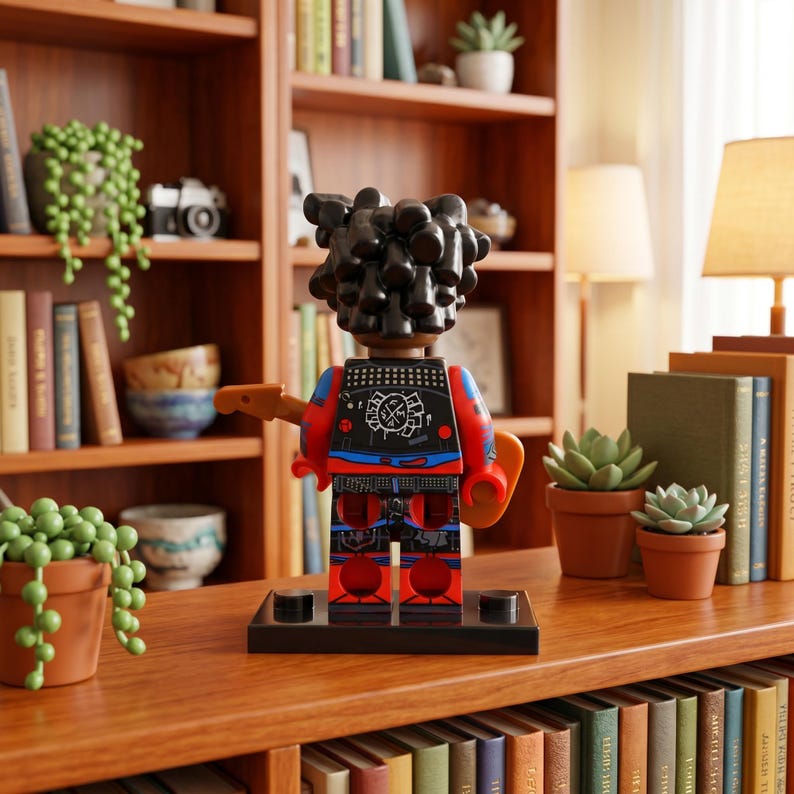 May include: A plastic toy figure with dark, textured hair, wearing a black vest with white graphic details and red sleeves, standing on a black rectangular base. The figure holds a brown guitar-like accessory and features blue and red leg details.