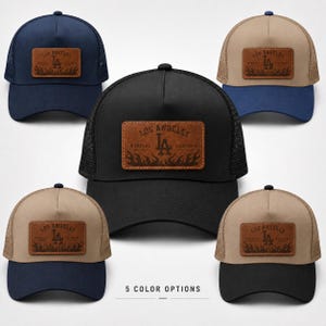 May include: Five trucker caps in various colour combinations, each featuring a rectangular brown leather patch with the text LOS ANGELES DODGERS CALIFORNIA and an LA logo above a flame graphic. The caps have mesh backs and solid front panels.