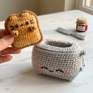 May include: A crocheted gray toaster with a smiling face and pink cheeks, paired with a tan crocheted toast slice featuring a cat face. A small crocheted jar labeled Yarn Preserve and a silver-colored butter knife are positioned nearby.