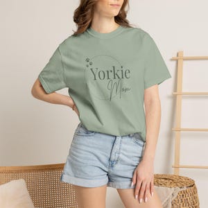Op de afbeelding: Een saliegroen T-shirt met korte mouwen en een donkergroene cirkelvormige afbeelding met de tekst Yorkie Mom en kleine pootafdrukken. Het shirt heeft een ronde hals en een relaxte pasvorm, gecombineerd met een lichtblauwe spijkerbroek.