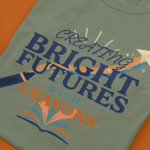 Pu&ograve; includere: Una t-shirt verde salvia con una grafica che riporta il testo "CREATING BRIGHT FUTURES WITH CRAYONS" in blu scuro e arancione. Il design include un'illustrazione di un aeroplanino di carta, un arcobaleno, un pastello e un libro aperto.