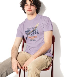 Pu&ograve; includere: Una t-shirt a maniche corte color viola chiaro con un design grafico che riporta il testo "CREATING BRIGHT FUTURES WITH CRAYONS" in blu e arancione, accompagnato dalle illustrazioni di un pastello, un aeroplanino di carta e un libro aperto.