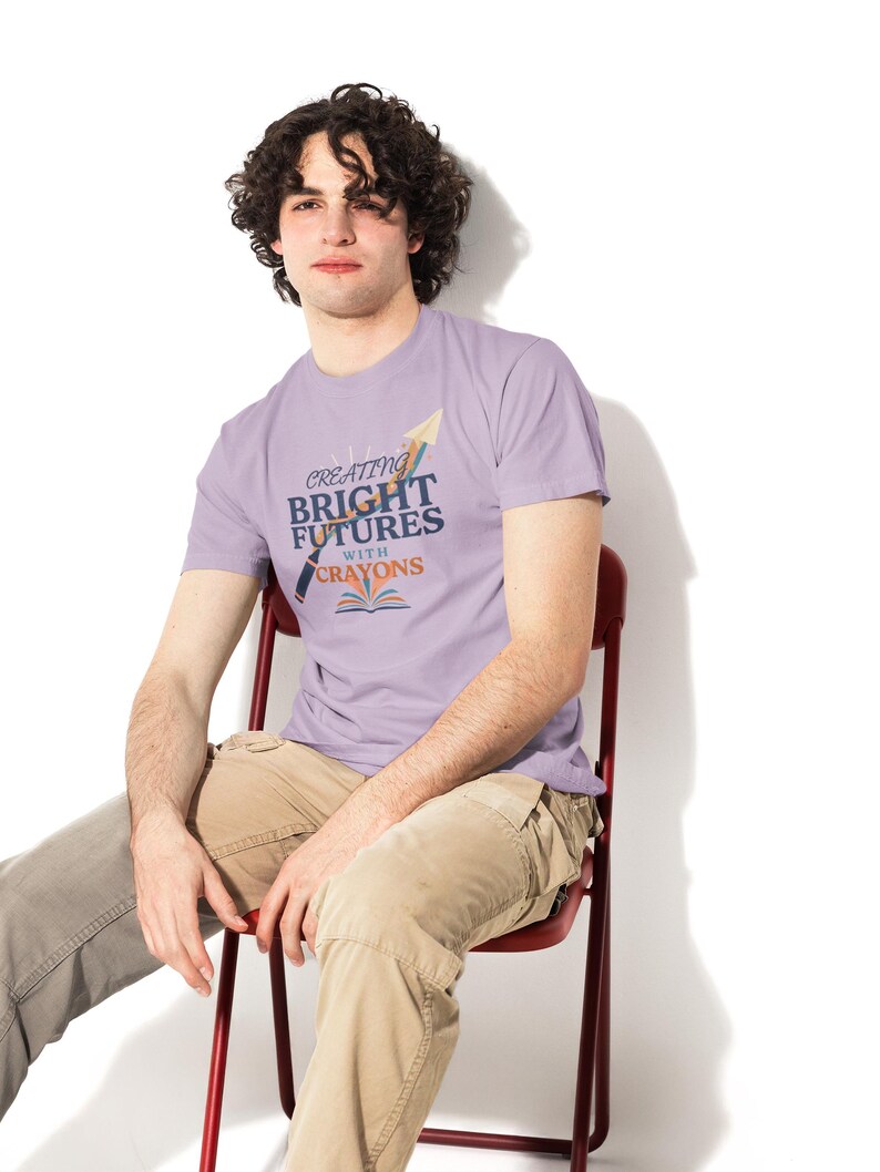 Pu&ograve; includere: Una t-shirt a maniche corte color viola chiaro con un design grafico che riporta il testo "CREATING BRIGHT FUTURES WITH CRAYONS" in blu e arancione, accompagnato dalle illustrazioni di un pastello, un aeroplanino di carta e un libro aperto.
