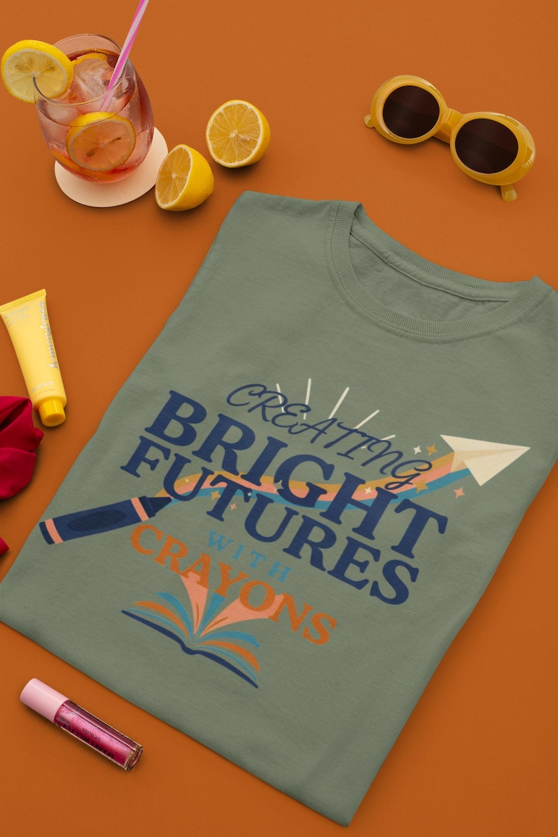 Pu&ograve; includere: Una t-shirt verde salvia con una grafica che riporta il testo "CREATING BRIGHT FUTURES WITH CRAYONS" in blu scuro e arancione. Il design include un'illustrazione di un aeroplanino di carta, un arcobaleno, un pastello e un libro aperto.