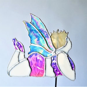 May include: A stained glass fairy figurine with iridescent blue wings, a white body, and pink and purple accents. The fairy is lying on its side with its head resting on its hand.