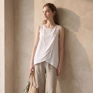 May include: A sleeveless white linen top with a diagonal overlapping front panel secured by three buttons at the shoulder, paired with straight-leg beige linen trousers, tan leather slide sandals, and a woven straw tote bag with leather handles.