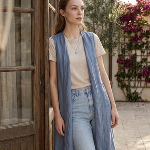 Puede incluir: Un chaleco largo azul sin mangas sobre una camiseta beige de manga corta y unos vaqueros azul claro. El chaleco presenta un diseño sencillo de frente abierto con un dobladillo recto que cae por debajo de las caderas y un collar dorado.