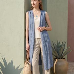 Könnte beinhalten: Eine ärmellose, knielange blaue Weste über einem cremefarbenen Tanktop und einer beigen Hose. Das Outfit umfasst eine goldene Halskette mit blauem Anhänger, eine goldene Armbanduhr und eine geflochtene Strohtasche mit braunen Lederhenkeln.