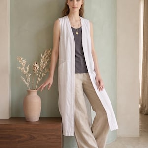 May include: A sleeveless white long vest worn over a dark gray tank top and loose-fitting beige trousers. The vest features a V-neckline, a long open front, and two visible side pockets. A gold pendant necklace is worn around the neck.