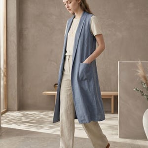 Könnte beinhalten: Eine lange, ärmellose blaue Weste mit Schalkragen und zwei aufgesetzten Vordertaschen, getragen über einem beigen Kurzarm-Oberteil und einer passenden beigen Hose mit weitem Bein, kombiniert mit braunen geflochtenen Sandalen und einer goldenen Kette.