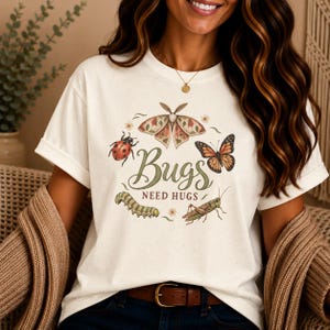 May include: A cream-colored t-shirt featuring a graphic print of various insects including a moth, a ladybug, a butterfly, a caterpillar, and a grasshopper. The text "Bugs NEED HUGS" is printed in the center in a decorative green and brown font.