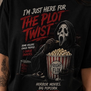 May include: A black t-shirt featuring a graphic of a masked figure in a black hooded robe eating popcorn from a bucket labeled FRIGHT FLIX. Text reads: I'M JUST HERE FOR THE PLOT TWIST, SOME KILLERS CHASE PEOPLE. I CHASE SNACKS, and HORROR MOVIES. BIG POPCORN. ZERO REGRETS.