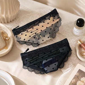Puede incluir: Dos estuches de malla negra semitransparente con un patrón de pequeños corazones negros y ribete de encaje negro en los bordes. El estuche superior contiene varias brochas de maquillaje, mientras que el inferior guarda un pequeño recipiente azul y negro.