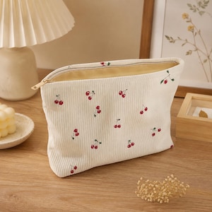 Può includere: Una pochette con cerniera in velluto a coste color crema caratterizzata da un motivo ripetuto di piccole ciliegie rosse con gambi verdi. La pochette presenta una chiusura a zip in metallo dorato e una fodera interna chiara visibile.