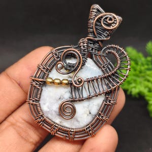 Könnte beinhalten: Ein Anhänger mit einem weißen Stein mit schwarzen Einschlüssen, der in kunstvoll gewickelten Kupferdraht gefasst ist. Das Design umfasst dekorative Spiralen, eine Öse an der Oberseite und drei kleine goldfarbene Metallperlen.
