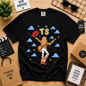 May include: A black t-shirt featuring a graphic of a person with long blonde hair wearing a yellow plaid shirt, cow-print pants, and red cowboy boots. The person holds a guitar and a red hat, surrounded by blue clouds, yellow stars, and the text TS.