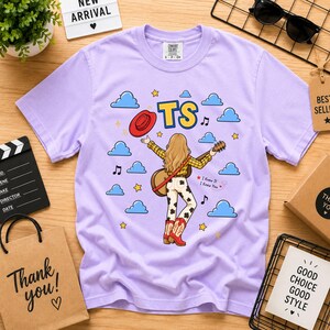 May include: A light purple short-sleeve t-shirt featuring a graphic of a person with long blonde hair playing a guitar, wearing a yellow plaid shirt and cow-print pants. The design includes a red hat, blue clouds, yellow stars, music notes, and the text TS.