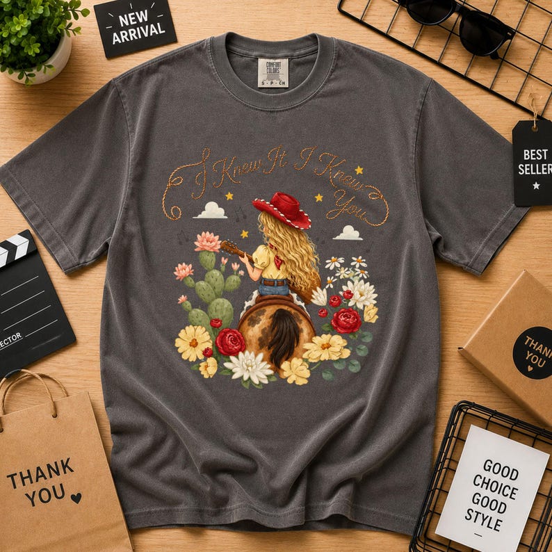 Puede incluir: Una camiseta gris carb&oacute;n con un gr&aacute;fico de una persona rubia con sombrero de vaquero rojo tocando la guitarra sobre un caballo. El dise&ntilde;o incluye cactus, flores coloridas, nubes, estrellas y el texto en cursiva I Knew It I Knew You.