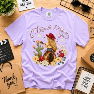 Puede incluir: Una camiseta de manga corta de color morado claro con un gr&aacute;fico de una persona de pelo largo rubio con un sombrero de vaquero rojo, tocando la guitarra sobre un caballo. El texto "I Knew It I Knew You" est&aacute; impreso encima, rodeado de cactus y flores.