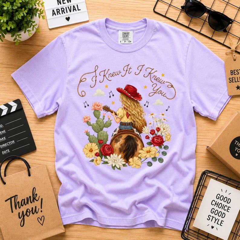 Puede incluir: Una camiseta de manga corta de color morado claro con un gr&aacute;fico de una persona de pelo largo rubio con un sombrero de vaquero rojo, tocando la guitarra sobre un caballo. El texto "I Knew It I Knew You" est&aacute; impreso encima, rodeado de cactus y flores.