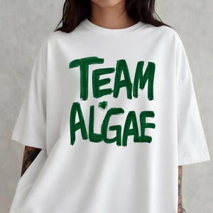 May include: A white short-sleeved t-shirt featuring the text "TEAM ALGAE" printed in a bold, dark green, brush-stroke style font across the chest. The garment has a crew neckline and a relaxed, loose-fitting silhouette with short sleeves.