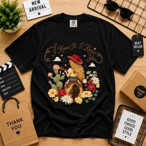 Puede incluir: Una camiseta negra de manga corta con el gr&aacute;fico de una persona de pelo largo rubio con sombrero de vaquero rojo, tocando la guitarra sentada en una silla de montar. El texto "I Knew It I Knew You" aparece impreso arriba, rodeado de cactus y flores.