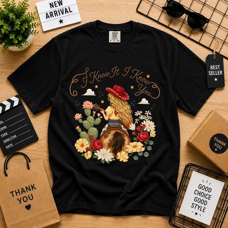 Puede incluir: Una camiseta negra de manga corta con el gr&aacute;fico de una persona de pelo largo rubio con sombrero de vaquero rojo, tocando la guitarra sentada en una silla de montar. El texto "I Knew It I Knew You" aparece impreso arriba, rodeado de cactus y flores.