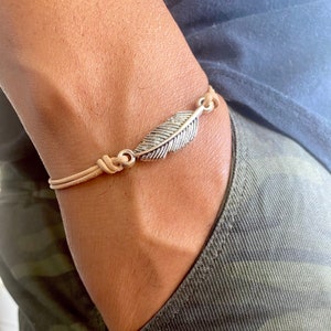 May include: A tan leather bracelet with a silver feather charm. The bracelet features a knotted design and a detailed feather pendant. The bracelet is a neutral color and is suitable for everyday wear.