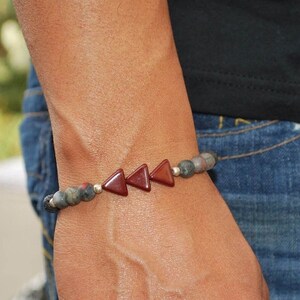 May include: A beaded bracelet featuring a series of dark red triangular stones interspersed with gray and green beads. The bracelet is strung on an elastic band and is worn on a wrist.