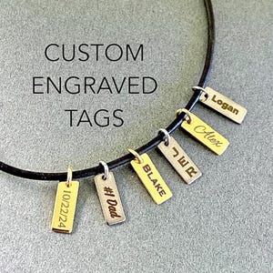 Puede incluir: Un collar de cuero negro con siete etiquetas rectangulares plateadas y doradas. Las etiquetas están grabadas con los nombres "Logan", "Alex", "JWR", "Blake", "#1 Dad" y "10/22/24".