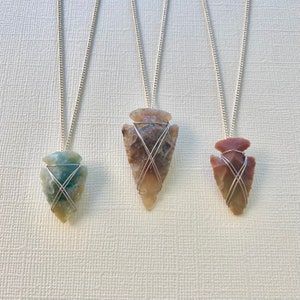 Peut inclure: Trois colliers en chaîne argent avec des pendentifs en forme de pointe de flèche. Les pointes de flèche sont faites de différents types de pierres : verte, brune et rouge.